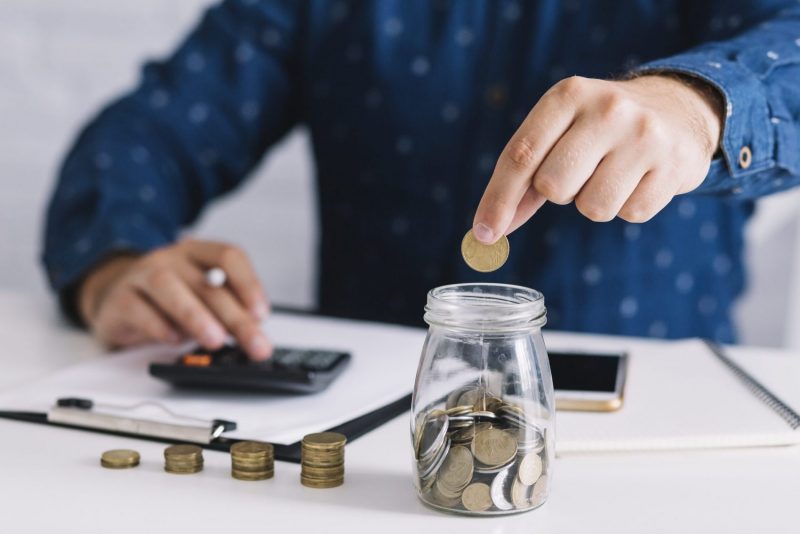 close-up-of-man-s-hand-putting-coin-in-jar-using-calculator